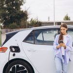 Eine junge Frau mit Smartphone in der Hand lehnt an ihrem E-Auto das gerade an einer Ladesäule geladen wird.