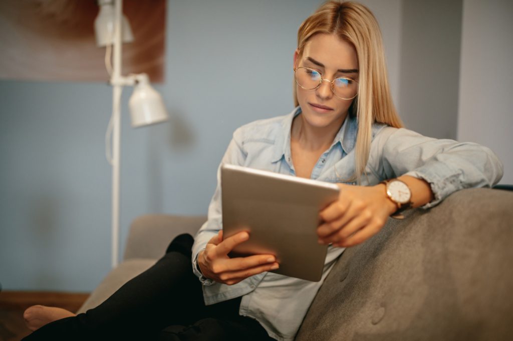 Eine junge Frau sitzt lässig auf einem Sofa und liest auf einem Tablet.