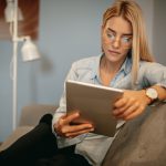 Eine junge Frau sitzt lässig auf einem Sofa und liest auf einem Tablet.