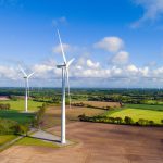 Mehrere Windräder stehen auf Feldern vor einem blauen Himmel mit leichten Wolken.