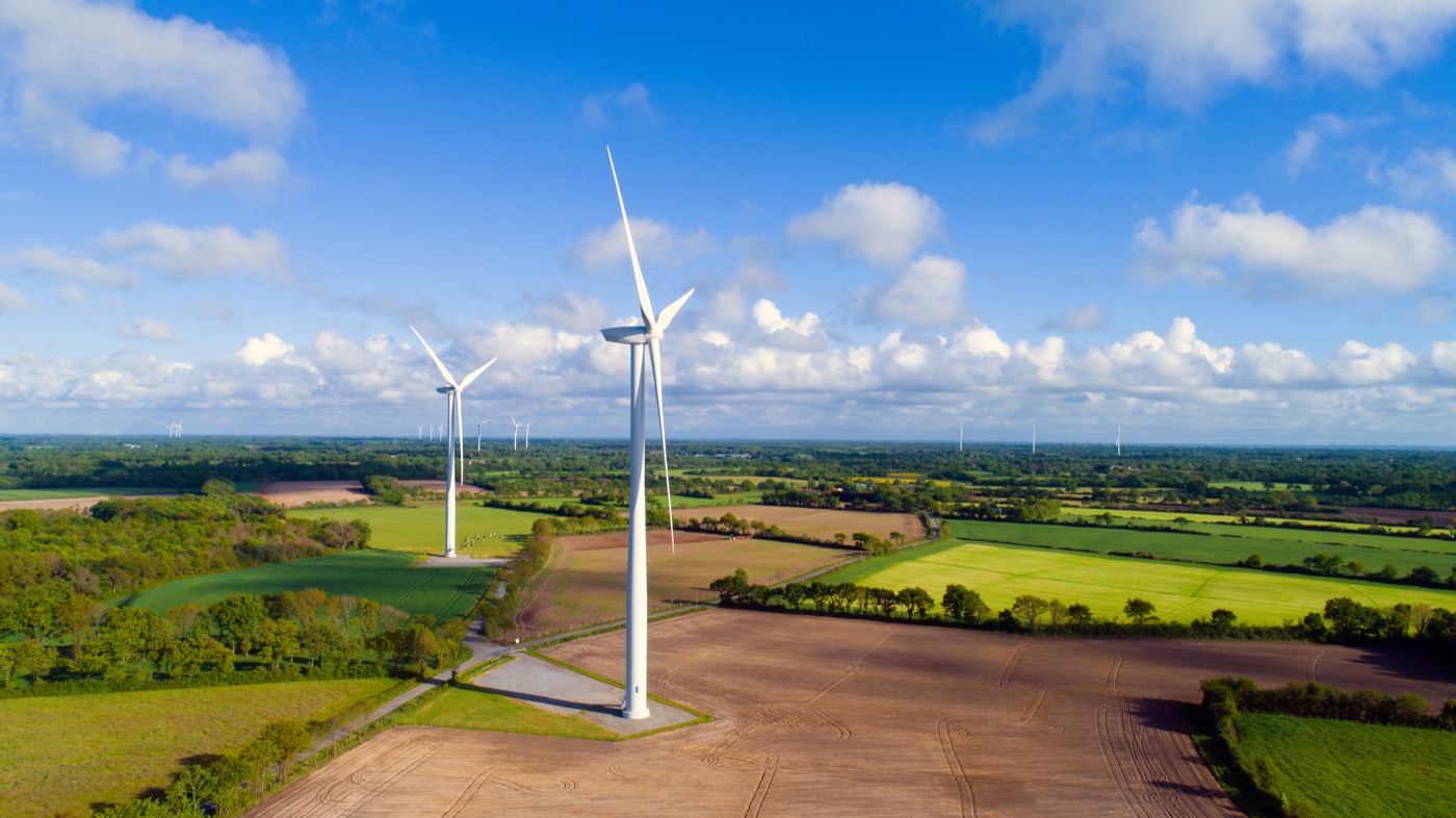 Mehrere Windräder stehen auf Feldern vor einem blauen Himmel mit leichten Wolken.