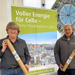 Frau Lyß (Leitung Marketing) und Herr Edathy (Geschäftsführer) stehen vor einem Rollup mit dem Celler Wimmelbild. Sie halten Papprollen in den Händen in denen das Wimmelbild als Poster enthalten ist.