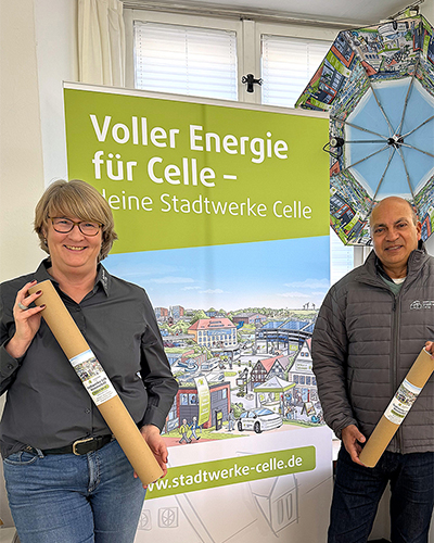 Frau Lyß (Leitung Marketing) und Herr Edathy (Geschäftsführer) stehen vor einem Rollup mit dem Celler Wimmelbild. Sie halten Papprollen in den Händen in denen das Wimmelbild als Poster enthalten ist.