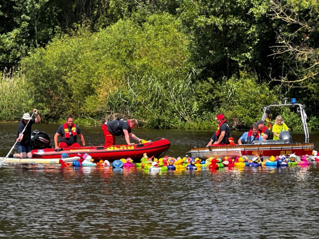 Zwei Boote des DLRG mit mehreren Mitarbeitenden des DLRG auf dem Wasser. Vor den Booten sind ganz viele Quietscheenten in verschiedenen Varianten im Wasser.