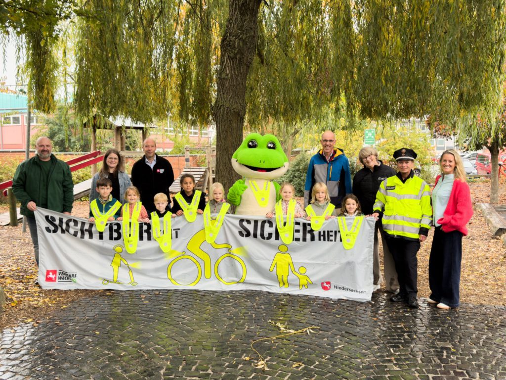 Mehrere Grundschulkinder halten ein Banner mit der Aufschrift "Sichtbar mit Sicherheit". Sie tragen Warnwesten und daneben stehen Mitarbeitende der Stadtwerke Celle, der Schule und von der Polizei.