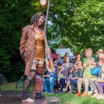 Ein fröhliches Publikum schaut sich draußen eine Vorführung an und lachen. Die Schausteller sind wie Clown geschminkt aber tragen sehr heruntergekommene Kleidung und stehen vor einem Puppentheater.