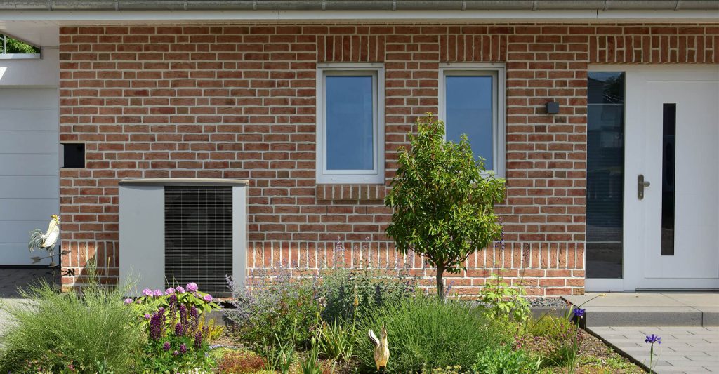 Eine Hauswand Haustür an der rechten Seite und auf der linken Seite eine Wärmepumpe. Im Vordergrund ein kleines Beet mit Stauden.